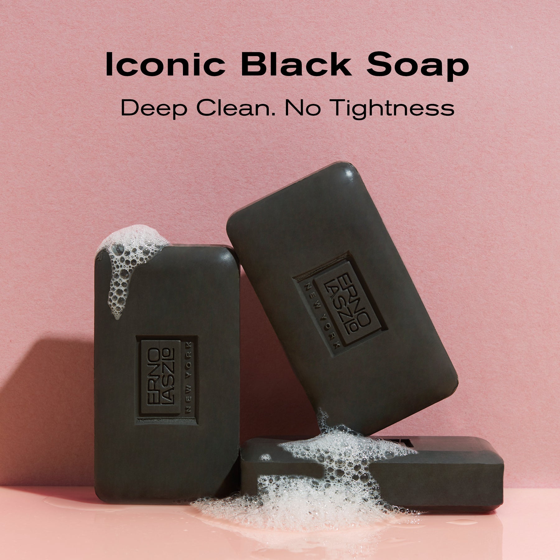 Three dark charcoal soap bars with white foam, arranged on a pink surface against a pink background. Text at the top reads 'Iconic Black Soap' and 'Deep Clean.
